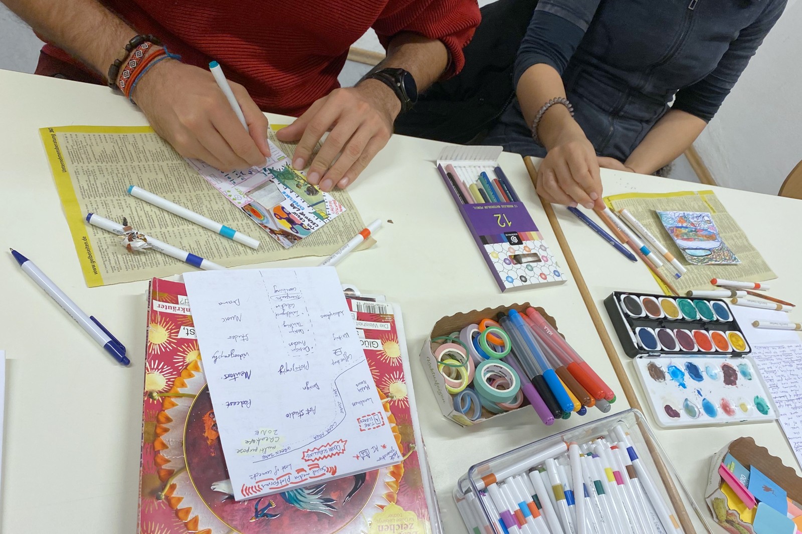 People writing in journals around a shared table at Open Page, paints and pages spread out