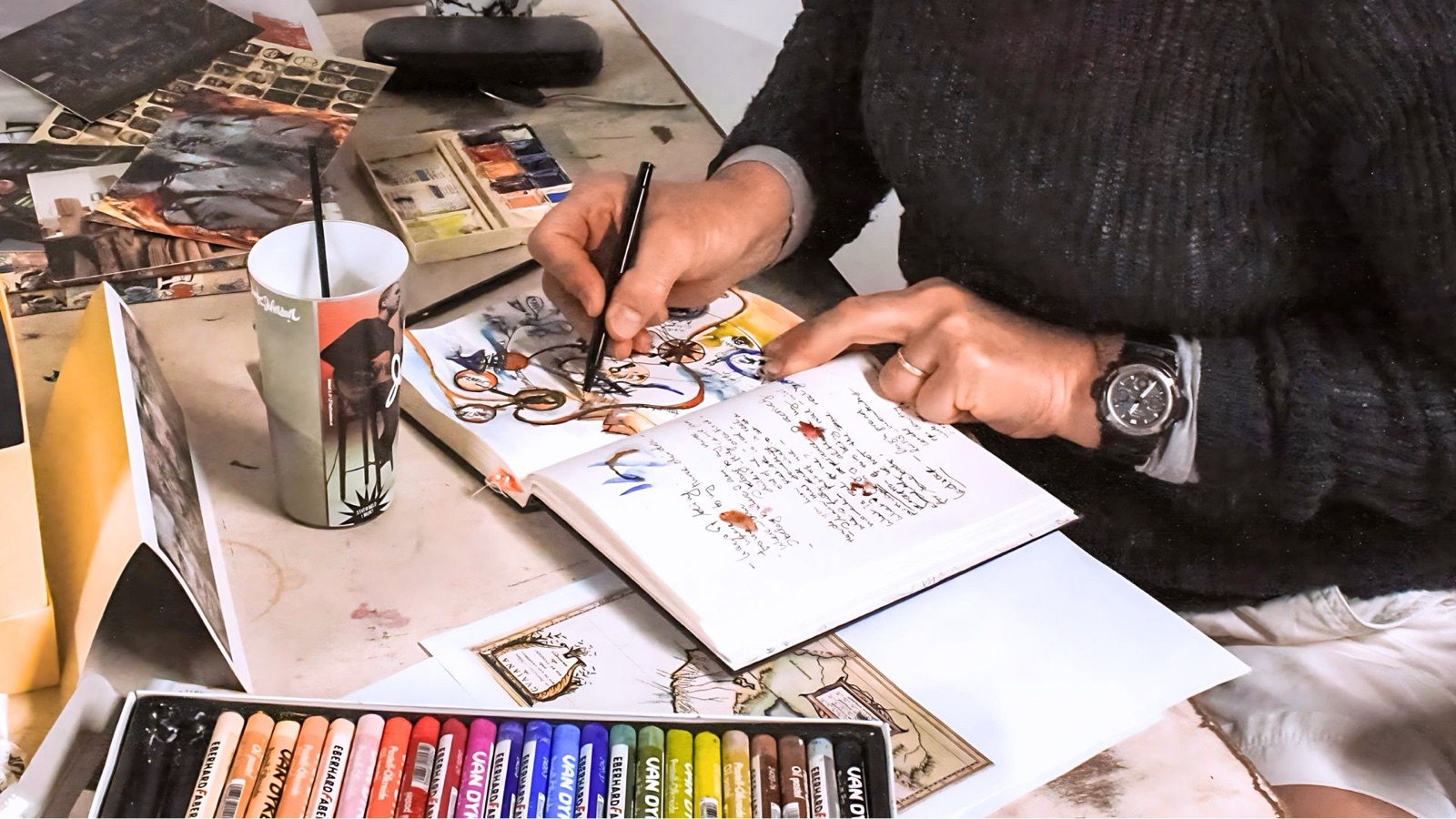 Hands working with oil pastels and a journal at a workshop table, art materials around
